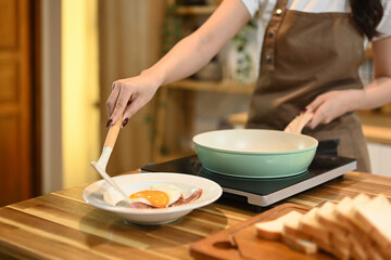 Young woman standing in a kitchen and serving fried egg and bacon onto a plate