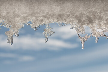 Melting snow hangs from the roof and shines in the sun, forming beautiful laces against the blue sky. Water drops create glare.