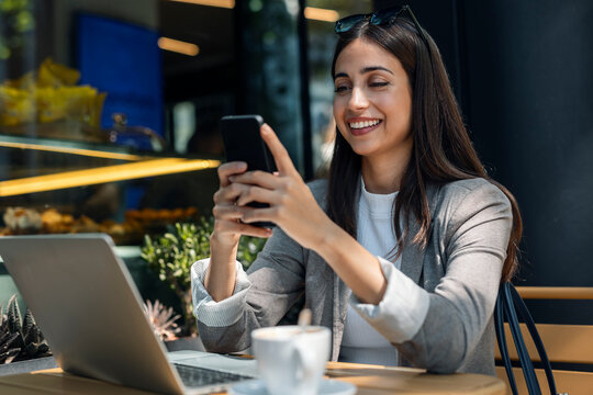 Happy young woman using smart phone and sitting with laptop in cafe