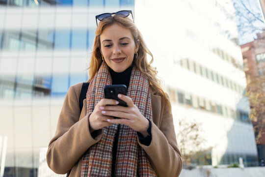 Smiling young woman in warm clothing using smart phone - Powered by Adobe