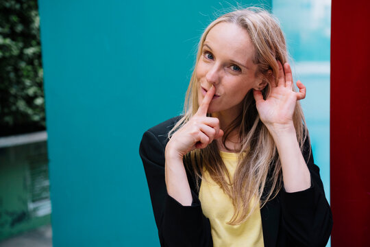 Happy businesswoman gesturing with finger on lips in front of blue wall
