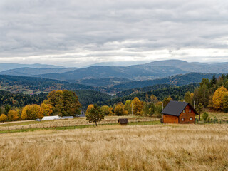 A wooden cabin sits on a grassy hill overlooking a valley. The valley is filled with rolling hills covered in lush green forests. A blue sky with fluffy white clouds can be seen in the distance.