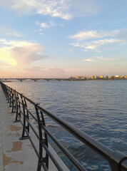 Fototapeta premium Embankment of Voronezh River is illuminated by rays of setting sun. There is road bridge inbackground. Metal barriers have been installed along embankment. Voronezh, Russia – June 17, 2024