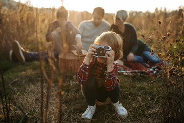 Fototapeta premium Portrait of family having picnic in autumn nature, eating homemade food in the middle of meadow. Young girl taking pictures with vintage camera.