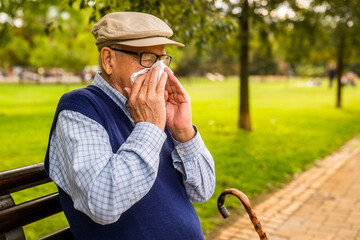 Portrait of senior man in park. He is sitting on bench and blowing nose.