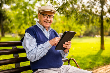 Fototapeta premium Portrait of happy senior man in park. He is sitting on bench and using digital tablet.