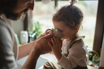 Father helping little daughter to drink warm tea from cup. Sick girl with cold is sitting on...