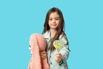 Cute little girl in stylish clothes with bunny toy and sweet lollipop on blue background