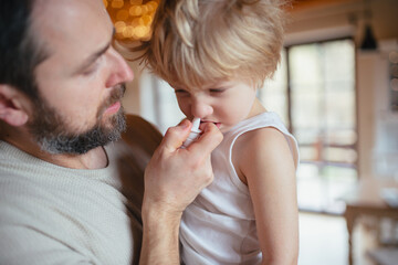 Sick boy with cold at home has stuffy nose. Father helping little son, using nasal spray in his...