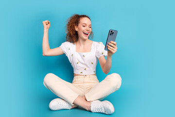 Full size photo of pretty young girl sit floor hold device winning excited wear trendy white outfit isolated on blue color background