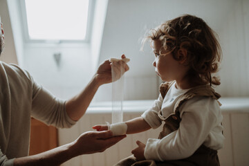 Father is bandaging his daughter's injured wrist in bathroom, concentrating while using a bandage.