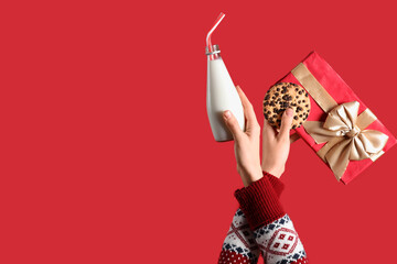 Hands holding Christmas gift, bottle of milk and cookie on red background