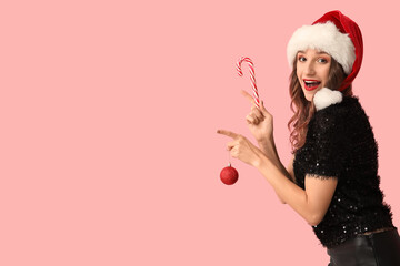 Young woman in Santa hat holding Christmas ball with candy cane and pointing at something on pink background