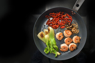 Frying pan with fried prawns and vegetables such as bok choy, red bell peppers and onions on a black background, cooking a healthy meal, low carb diet with protein, copy space, from above