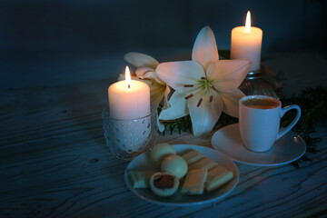 Two candles, white lily flowers, coffee cup and pralines, festive light against a dark blueish background, still life for Advent, Christmas and winter holidays, copy space