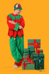 Handsome young man in elf's costume pointing at gift boxes on yellow background