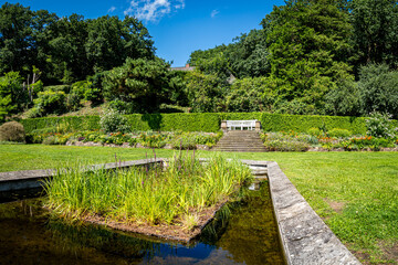 Surrounded by greenery and flower beds, the R&ouml;mischer Garten in Hamburg Blankenese offers a outdoor retreat featuring a reflecting pond and a white bench, perfect for enjoying peaceful tranquility.
