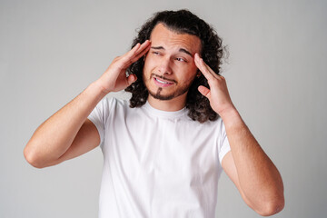 A young man with curly hair rubs his forehead in a casual indoor setting while expressing...