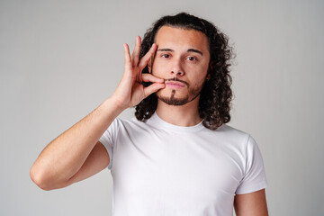 A young man with curly hair demonstrates a hand gesture in a plain white shirt against a neutral background