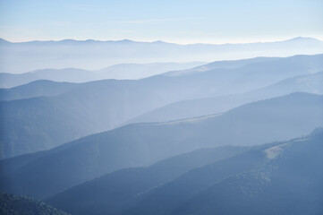 Fototapeta premium Soft blue hues of layered mountain ranges stretch into the distance under clear sky, creating tranquil and atmospheric landscape filled with gentle gradients and sense of vastness.