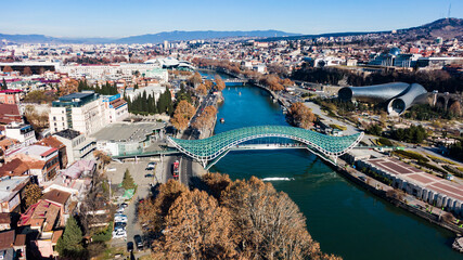 Aerial view of the bridge of peace spanning the kura river in central tbilisi, with the modern architecture of the music hall and the foothills of the caucasus mountains in the background © wifesun