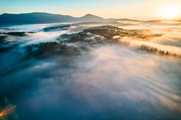 Stunning aerial view of sunrise over mountainous landscape, with thick fog blanketing valleys and hills. Sun's golden rays pierce through mist, casting serene glow over the entire scene.