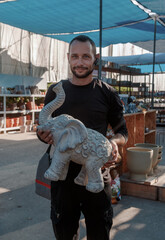 In an open-air outdoor market, a man proudly displays a elephant sculpture highlighting the artistry and craftsmanship present in the various decorative items around him. Eelephant statue. Marketplace