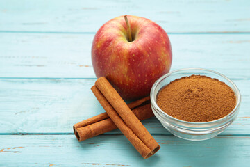 Red apple, cinnamon sticks and cinnamon in bowl on blue wooden background.