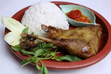 Traditional Indonesian fried chicken with fresh vegetables and chili sauce, served on a wooden plate, isolated on a white background, top view.