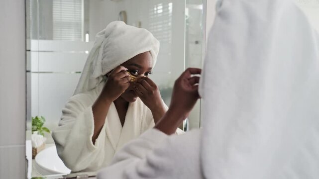 Over the shoulder of African American woman in white bathrobe applying patches under eyes during self-care treatment routine at home