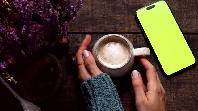Cozy flat lay of female holding warm coffee mug with green screen chroma key smartphone. Decorated with lavender flowers, chocolate chunks and chestnuts on dark rustic wooden table in autumn.