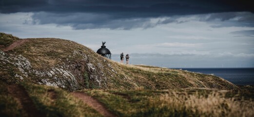 Hikers on a coastal path with a lighthouse and dramatic sky.