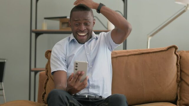 African Man Excited for Results on Phone while Sitting on Sofa at Home