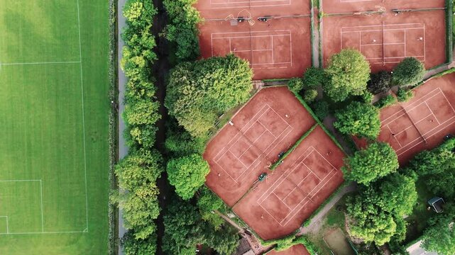 tennis courts with people playing, surrounded by trees, with a nearby road, and a soccer pitch