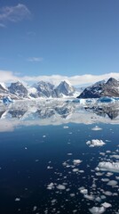 Serene Arctic image with iceberg in calm waters. Whites and blues contrast, with snowy mountains in backdrop. Clear sky reflects on water, evoking peacefulness and pristine beauty.