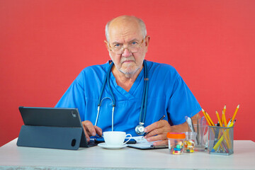 An elderly man, a doctor, in his office, at the table. Blue background.