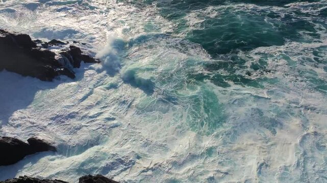 Drone slow-motion of foamy ocean waves breaking on the rocky shore of Seixo Branco on a sunny day