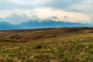 Stunning mountain range with snow-capped peaks overlooking rolling grassy hills. A picturesque scene capturing the beauty of nature, the transition from green meadows to high-altitude mountains 