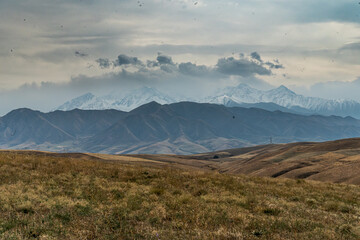 Expansive mountain view beneath dramatic sky. Dry, golden grasslands stretch towards snow-capped...