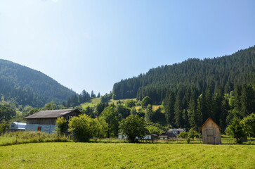 Scenic view of a traditional wooden houses with a dark roof in a lush green valley, surrounded by dense forested hills under clear blue skies. Carpathian Mountains, Ukraine