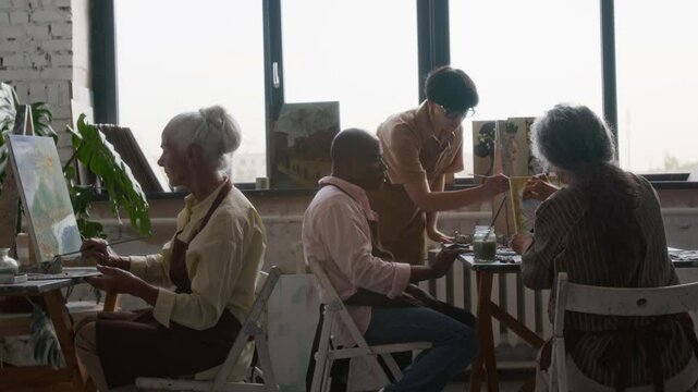 Female teacher helping senior man finish his picture, two women painting in art studio during workshop