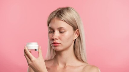 Young blonde woman posing at camera, opens jar and applies cream on face, isolated on pink studio background. Beauty skin care concept