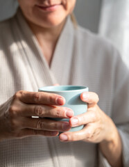 Mature caucasian woman is drinking tea or coffee from cup near window at home. Concept of domestic lifestyle. Cropped photo of modern happy middle aged lady wearing bathrobe at sunny day.