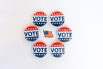 Vote buttons and American flag pin on white background. A collection of red, white, and blue "Vote" buttons arranged in a circular pattern around a small American flag pin symbolizing patriotic voting
