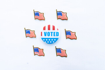 An "I Voted" button surrounded by six small American flag pins arranged in a circular pattern on a white background. The button features red, white, and blue colors with stars, representing the voting
