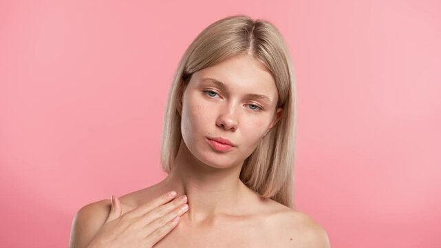 Young blonde woman posing at camera, touches neck and collarbone smooth skin, isolated on pink studio background. Beauty skin care concept