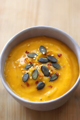 Bowl of creamy pumpkin and potato soup, with pumpkin seeds and chilli flakes toppings. Selective focus, wooden background.