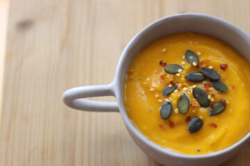 Bowl of creamy pumpkin and potato soup, with pumpkin seeds and chilli flakes toppings. Selective focus, wooden background.