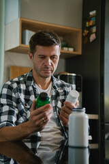 Man is sitting at table in kitchen looking at various bottles of medicine capsules. Person is taking pills or vitamins. Concept of supplements, medicine or health care