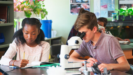 High School Students Looking Through Microscope And Writing Notes In Biology Class